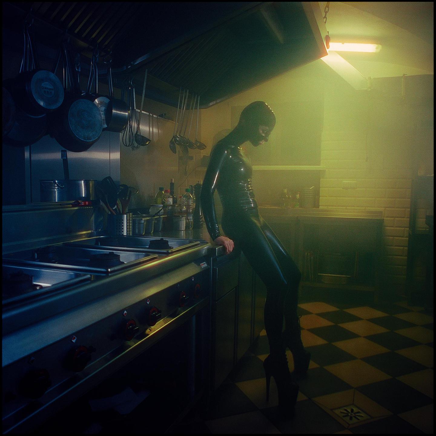 A person in a black latex suit and high heels leans on a counter in a commercial kitchen.