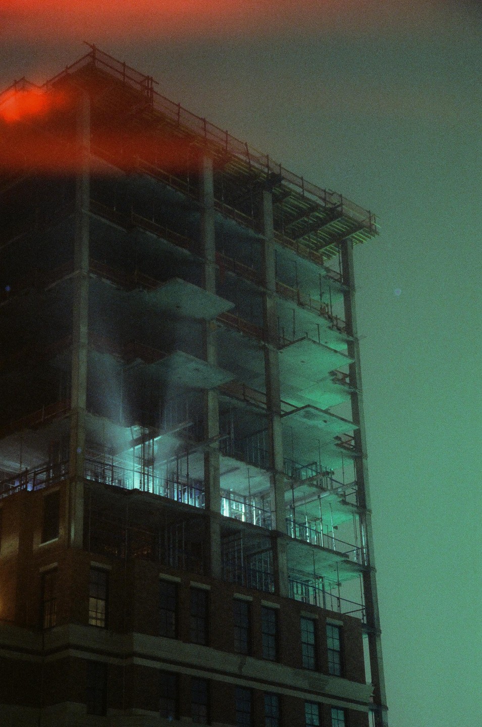 A tall building under construction is illuminated with green light at night, with a red light leak across the top.