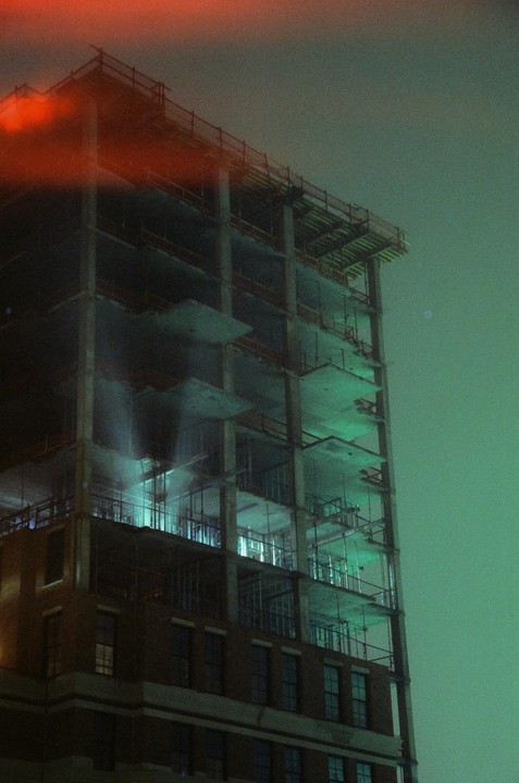 A tall building under construction is illuminated with green light at night, with a red light leak across the top.