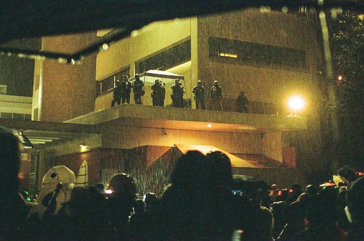 A group of people stand in the rain at night, looking up at a building where several armed men are visible on a balcony.