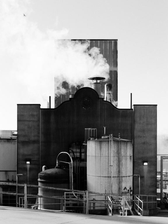 A large industrial building with smoke rising from its chimneys is shown in black and white.