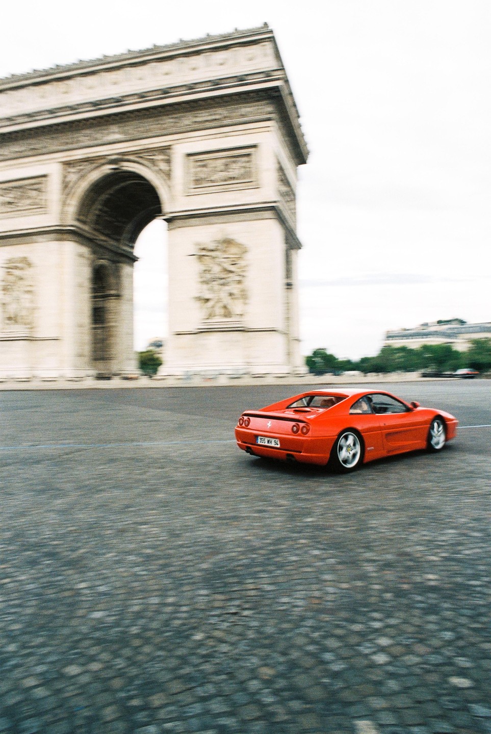 A red sports car drives on a cobblestone street with the Arc de Triomphe in the background.