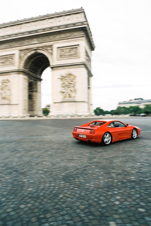 A red sports car drives on a cobblestone street with the Arc de Triomphe in the background.