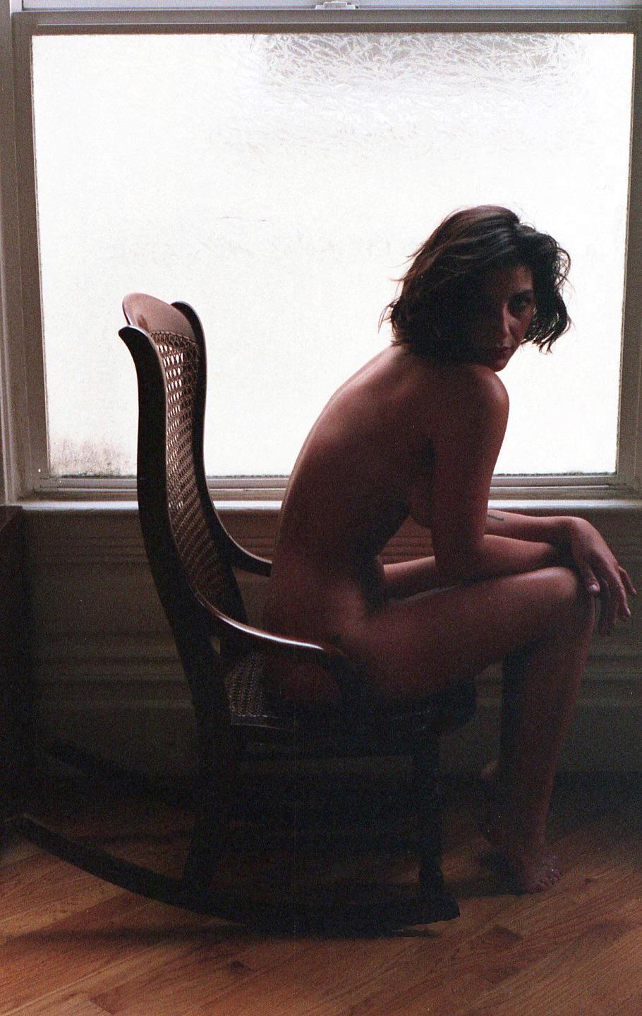 A woman sits nude in a rocking chair, looking over her shoulder at the viewer.