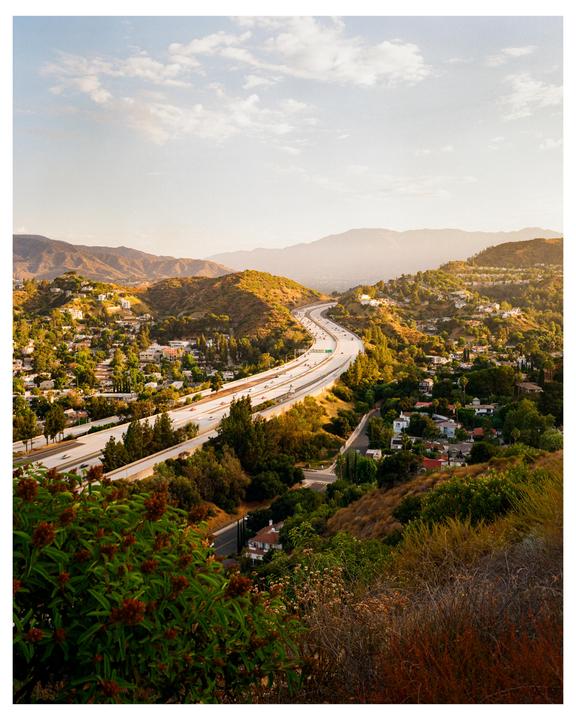 A freeway winds through a valley filled with houses and trees, with mountains in the distance under a hazy sky.
