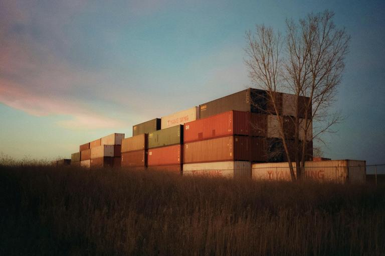 A stack of shipping containers sits behind a field of tall grass with a bare tree to the right under a blue and pink sky.