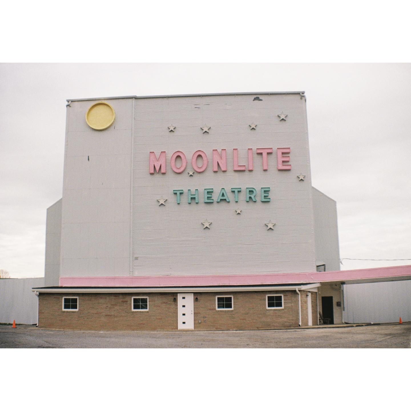 The Moonlite Theatre building has pink and green lettering on a white facade with a brick base.