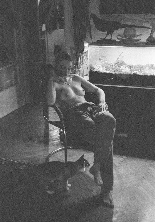A topless woman sits in a chair next to an aquarium while a cat walks on the floor in front of her.