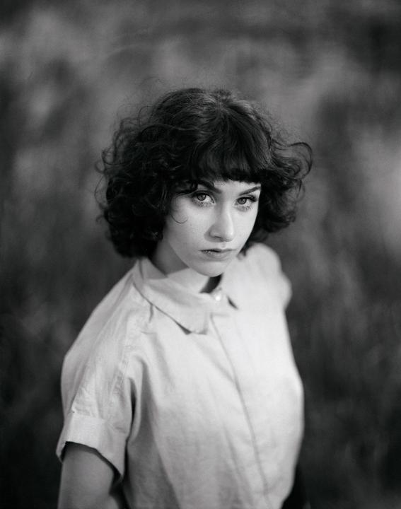 A woman with curly hair and bangs looks directly at the viewer.