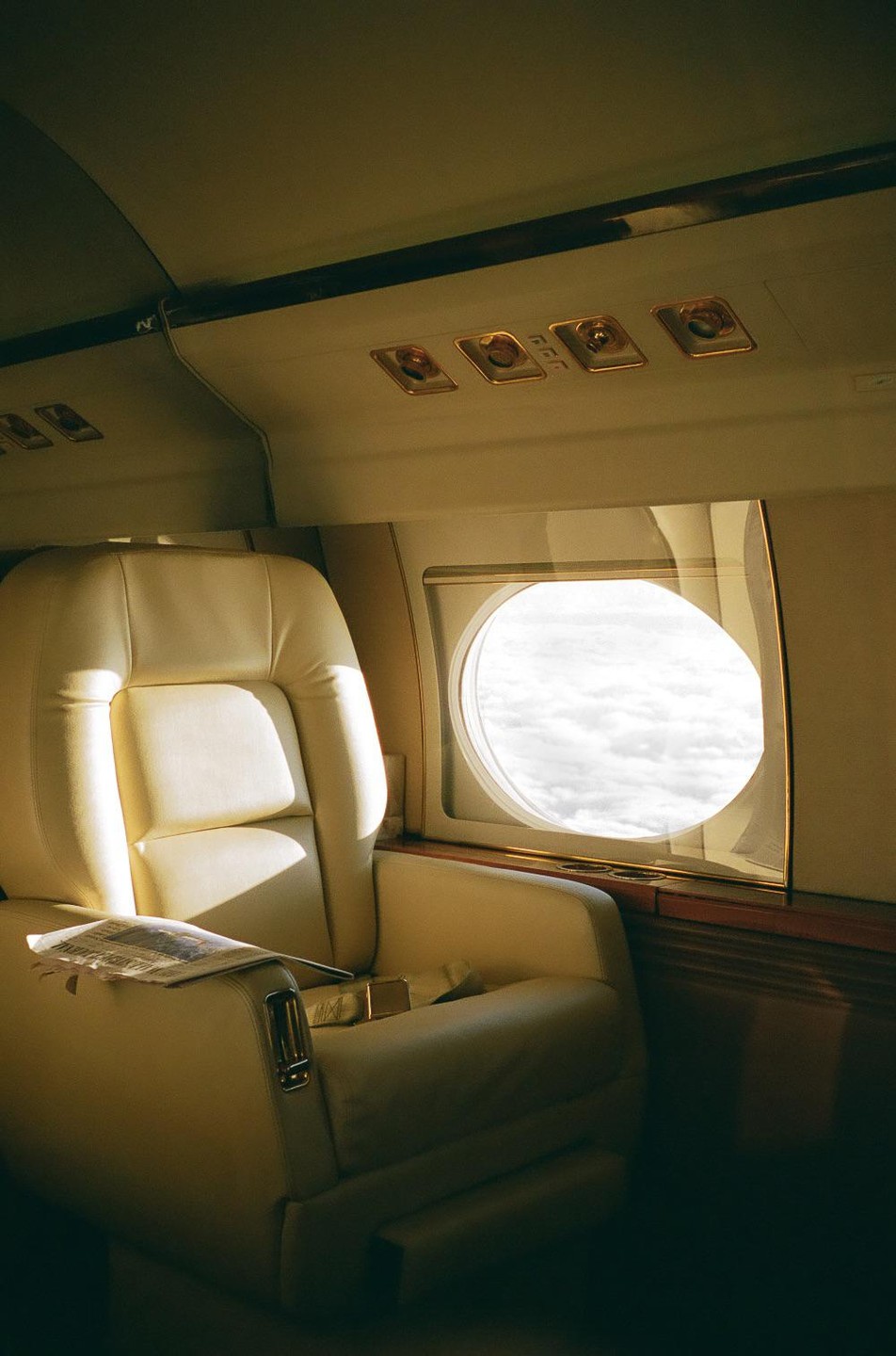 An empty cream-colored airplane seat is next to a window looking out at clouds.