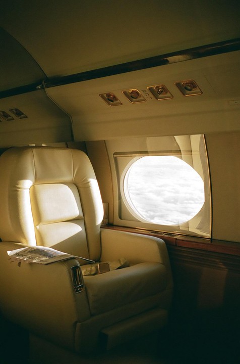 An empty cream-colored airplane seat is next to a window looking out at clouds.
