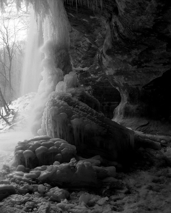 A waterfall cascades over a rock face covered in ice formations and icicles, with bare trees visible in the background.