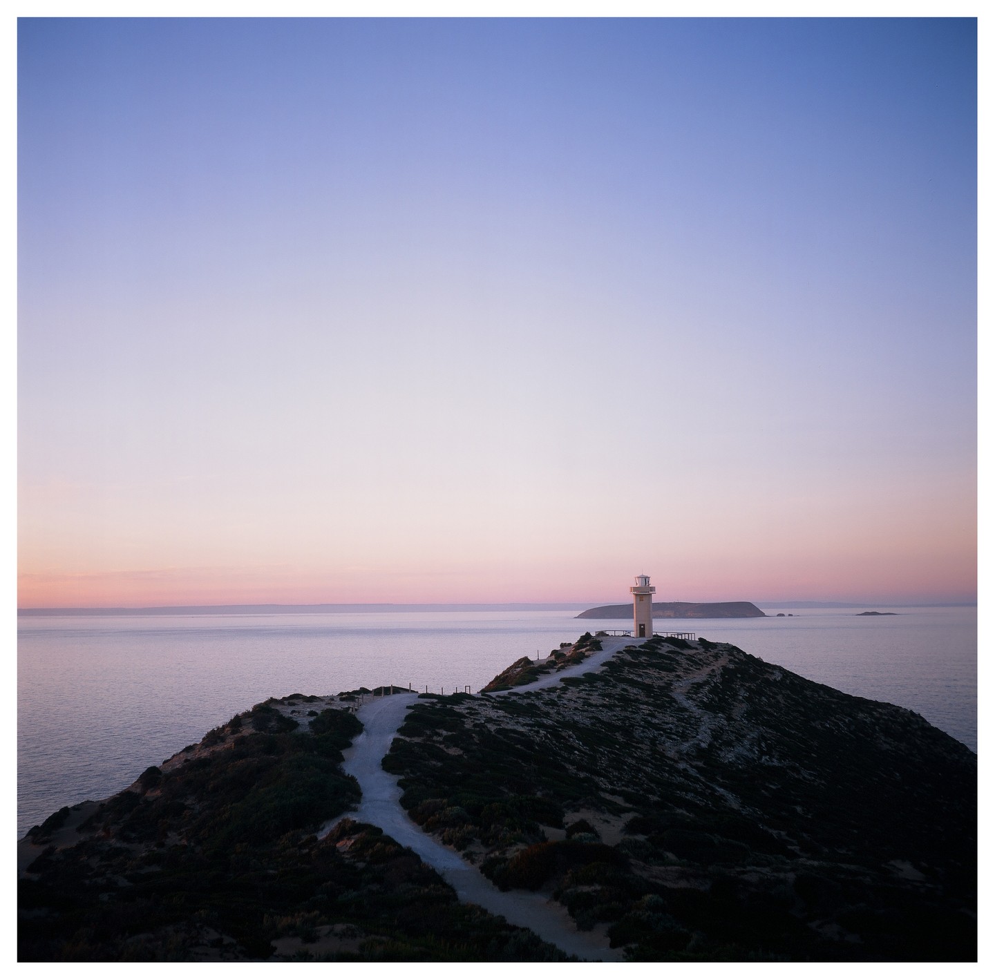 A lighthouse stands on a hill overlooking the ocean at dusk, with a path leading up to it.