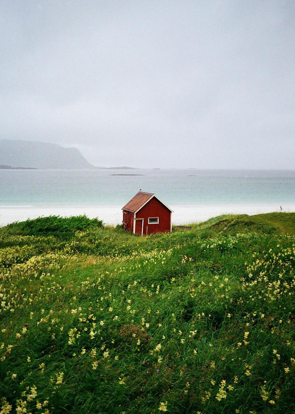 A red cabin sits on a grassy hill overlooking a white sand beach and the ocean under a cloudy sky.