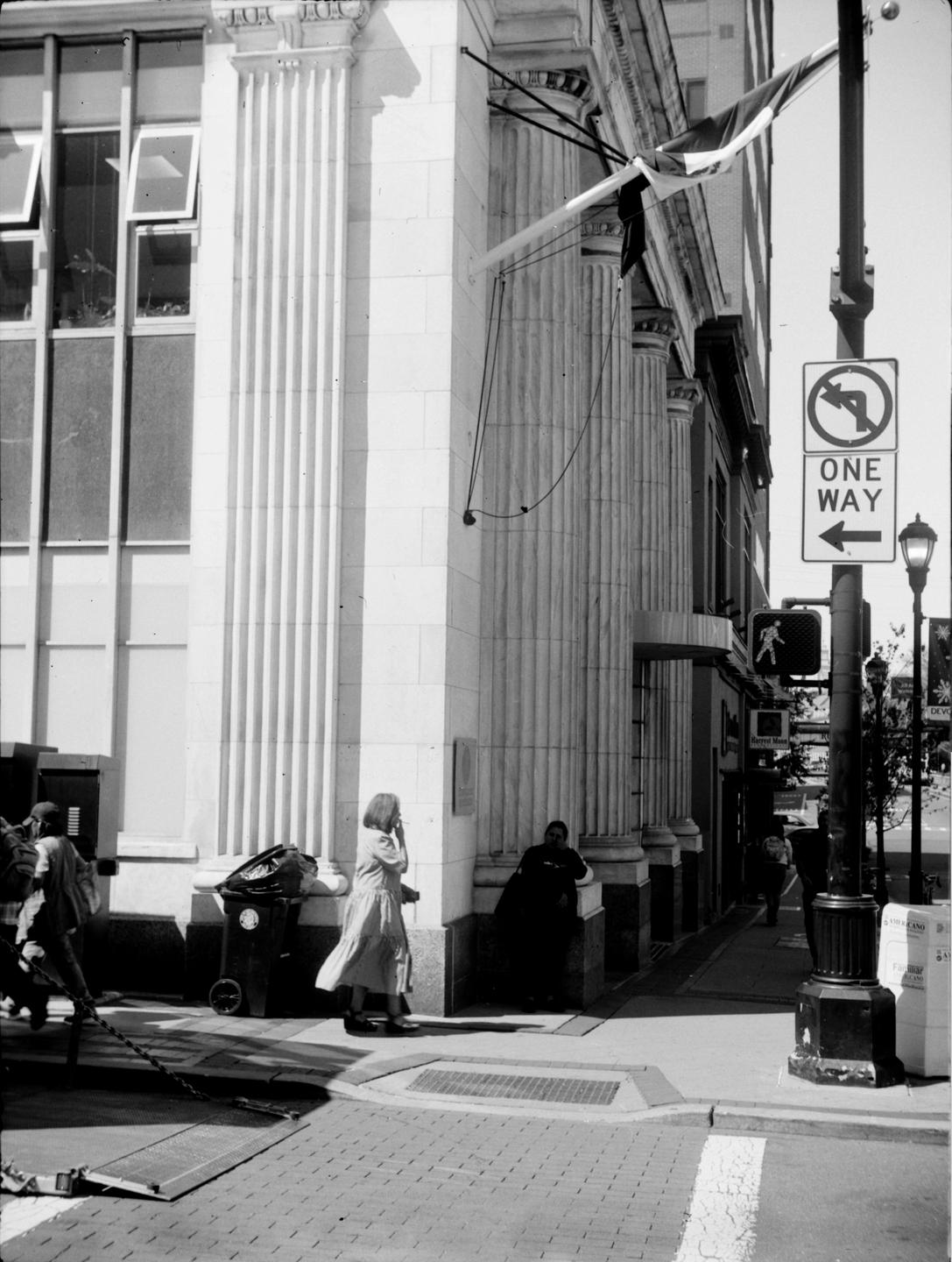 A woman walks past a building with large columns while another woman sits in the shadow of the building.
