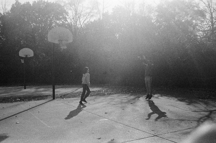 Two children play basketball on an outdoor court with trees in the background.