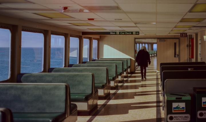 A woman walks down the aisle of a ferry with rows of seats and windows looking out onto the water.