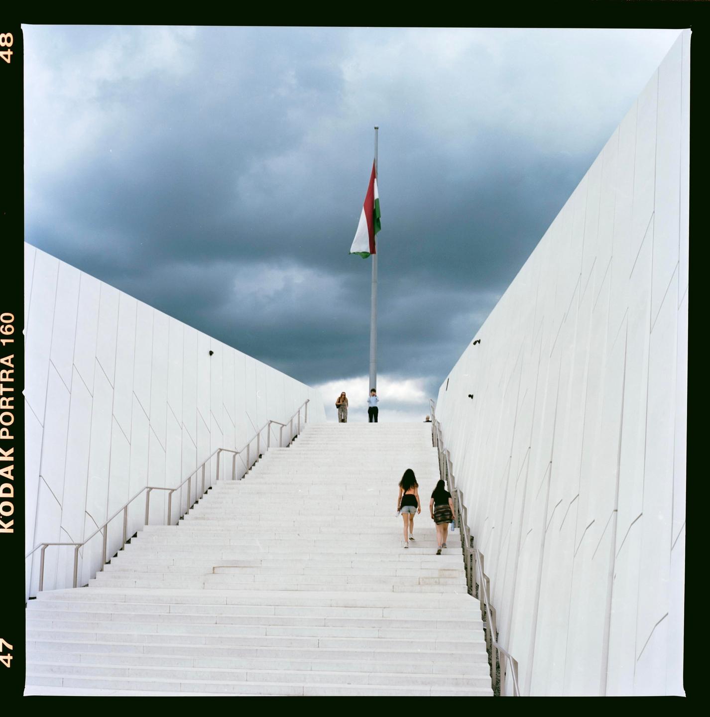 A Hungarian flag flies above a long, wide, white staircase with people walking up it under a cloudy sky.