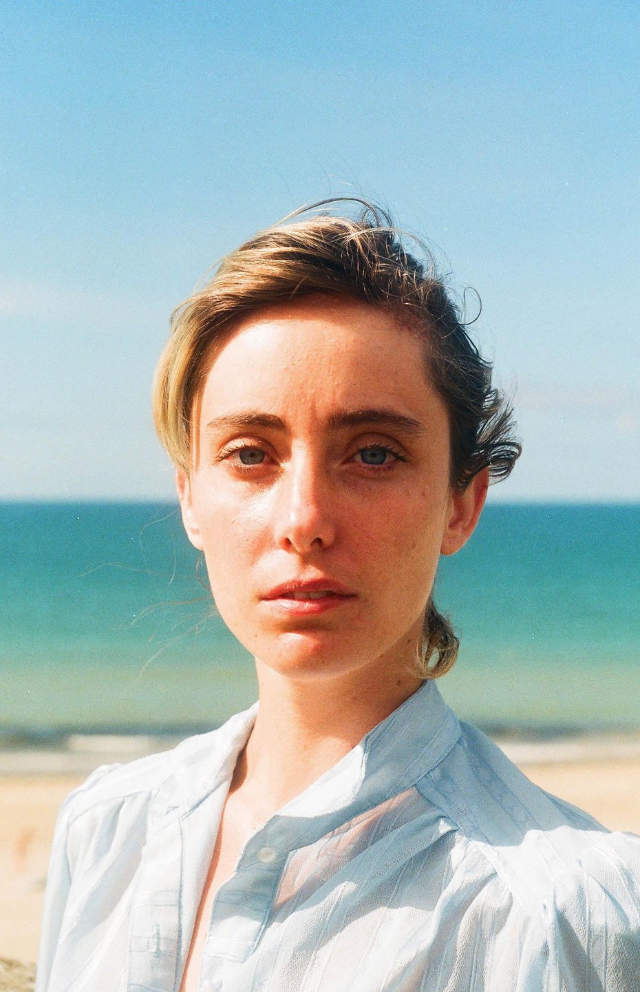 A woman with blonde hair and a light blue shirt looks directly at the viewer with the ocean and beach in the background.