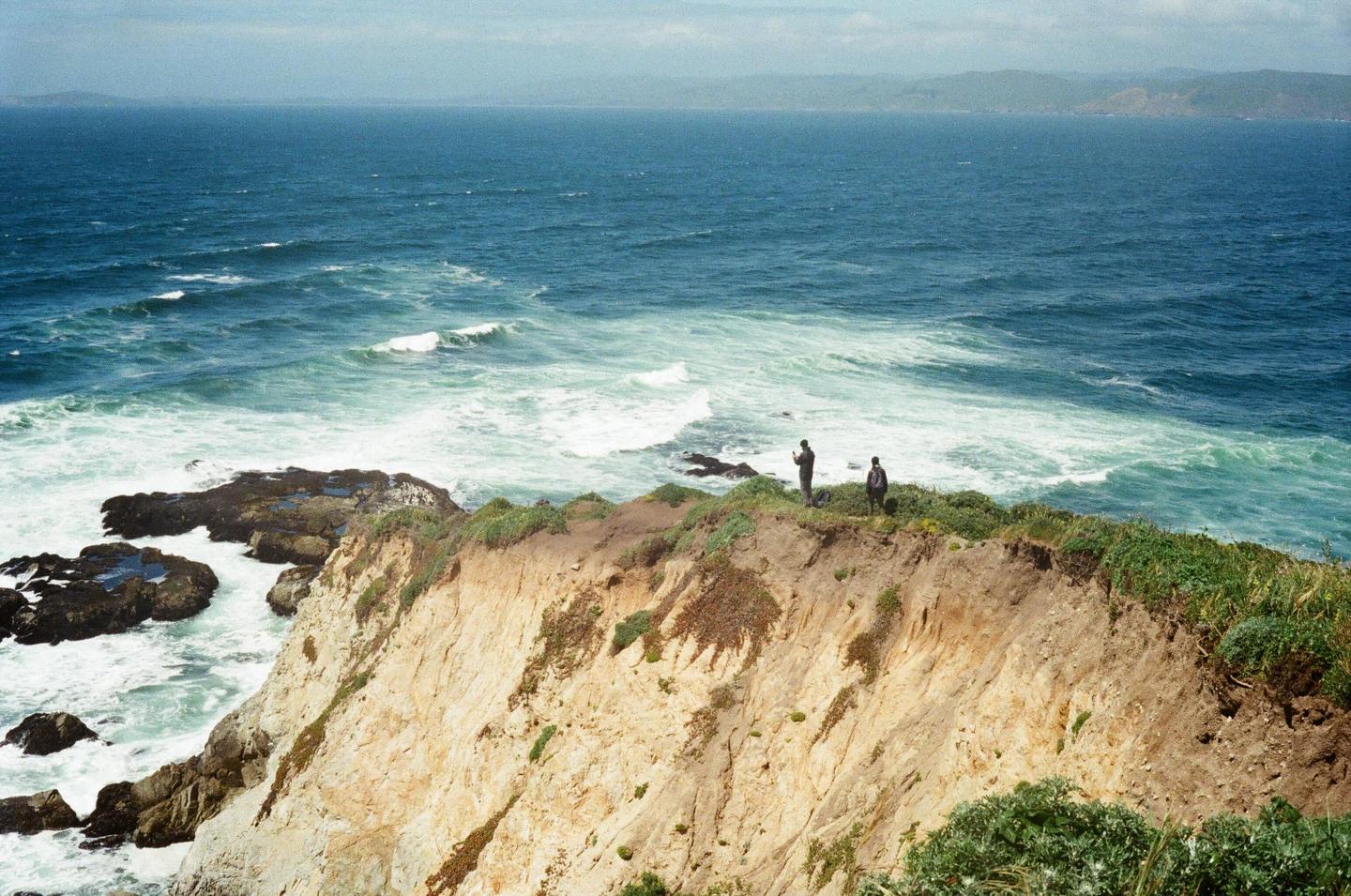 Two men stand on a cliff overlooking a vast ocean with waves crashing against the shore.