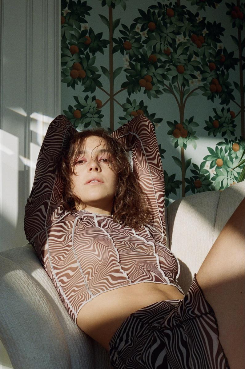 A woman with curly hair and a patterned top is reclining on a chair in a room with orange tree wallpaper.