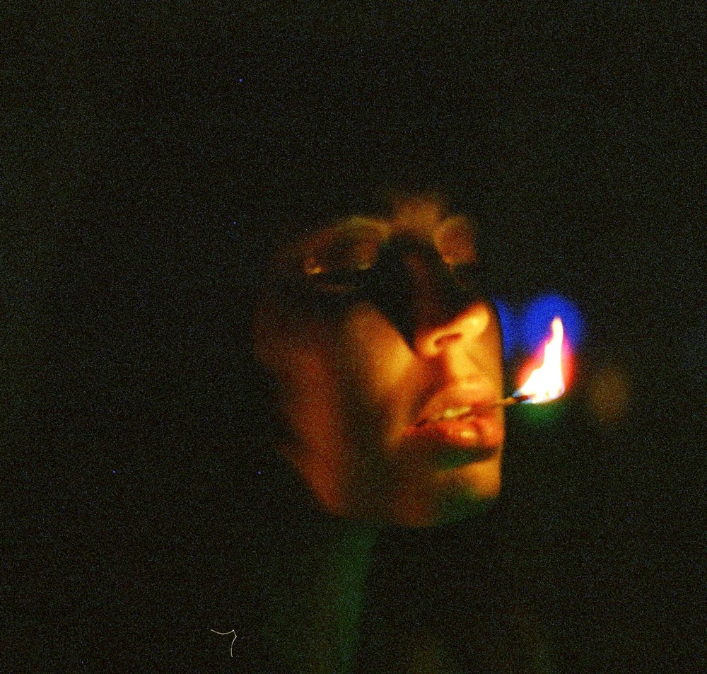 A person's face is illuminated by the flame of a match held in their mouth in a dark setting.