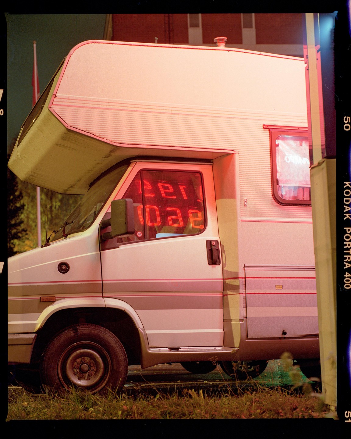 A white camper van is parked on grass next to a building with a red neon sign reflected in its window.