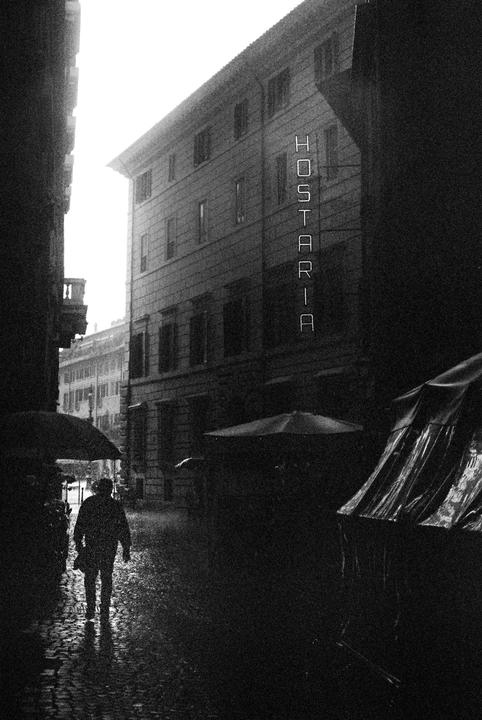 A person walks with an umbrella on a wet cobblestone street next to a building with a "HOSTARIA" sign.