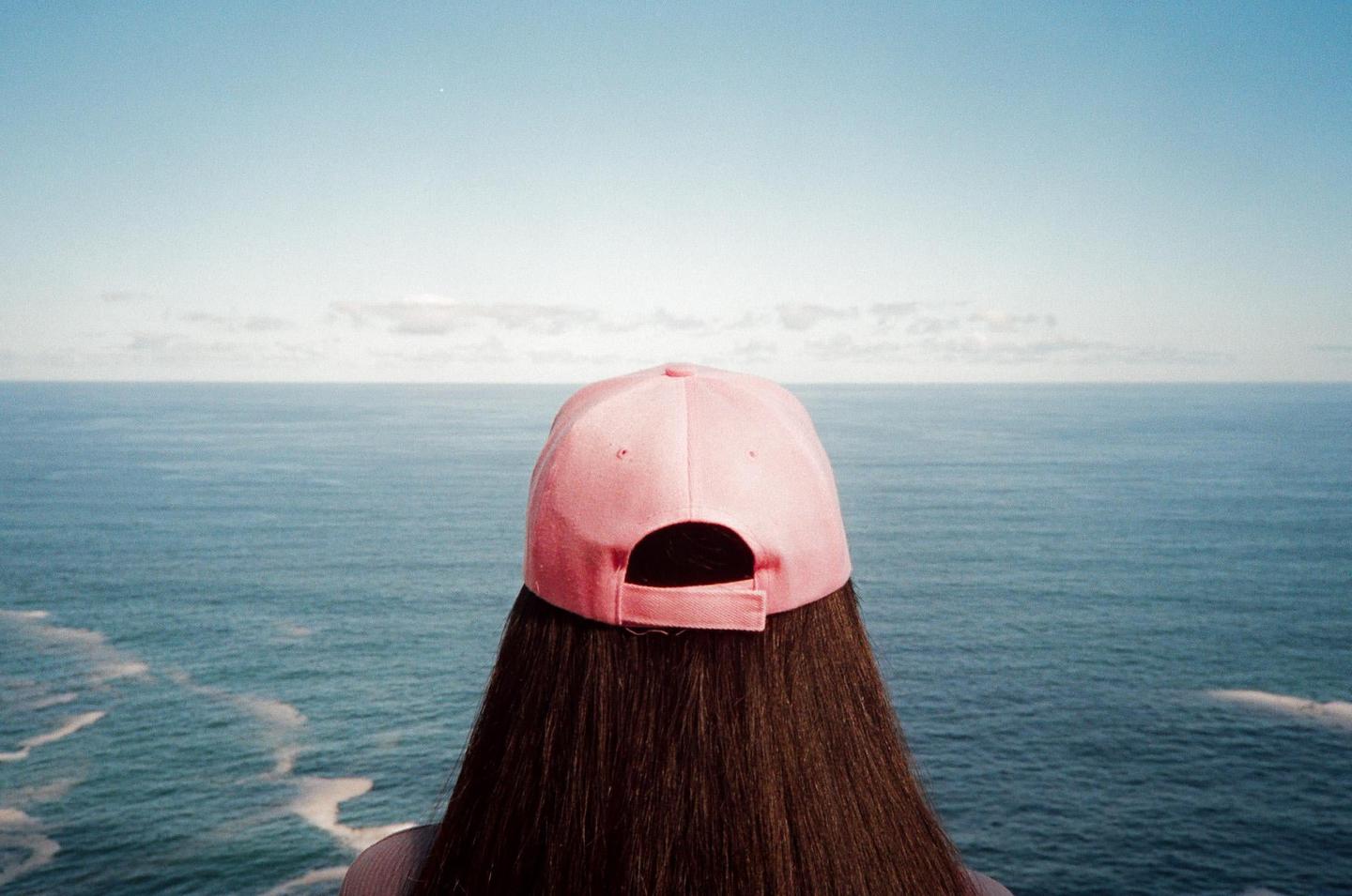 A person with long hair and a pink baseball cap looks out over the ocean under a clear sky.