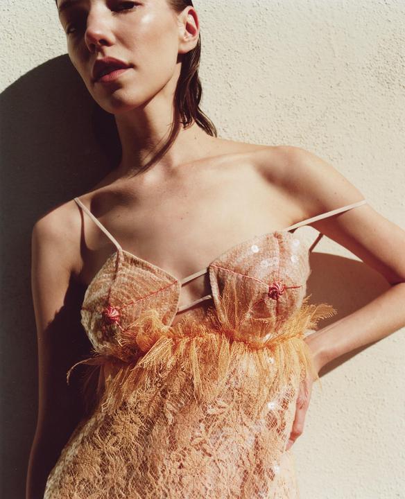 A woman with wet hair and a sequined, feathered dress stands against a wall in direct sunlight.