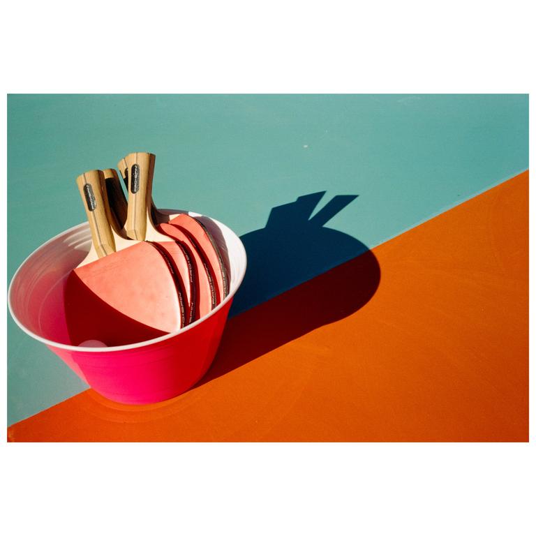 A pink bucket holds several table tennis paddles and a ball on a two-toned surface.
