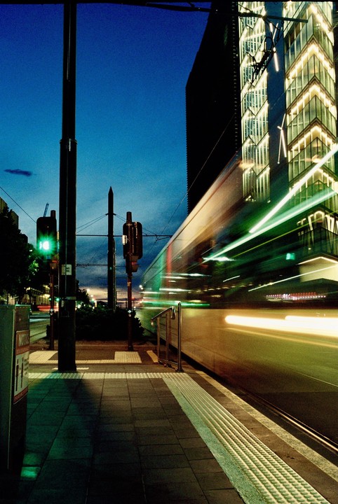 A tram blurs past a modern building with illuminated windows at dusk.