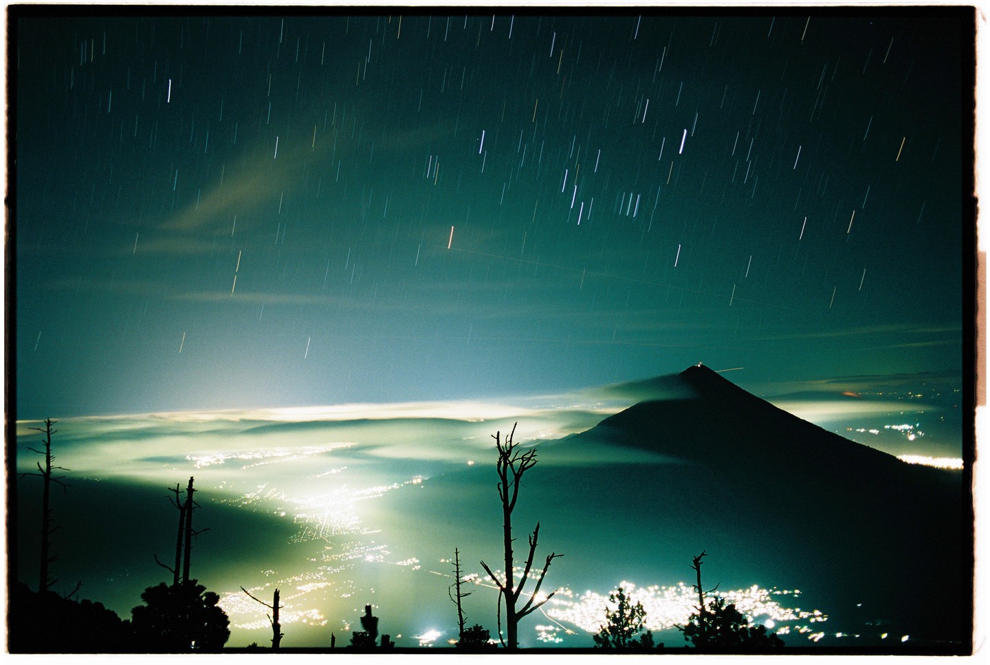 A long exposure photograph captures star trails above a volcano and a city illuminated at night.