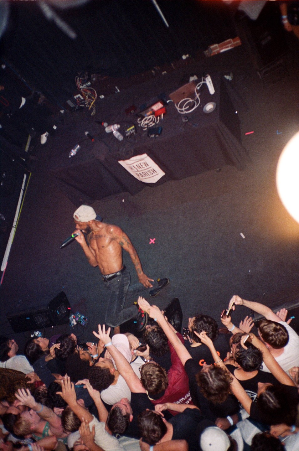A shirtless man with a microphone performs on stage for a crowd of people reaching up towards him.