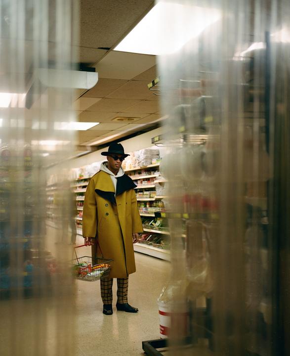 A man in a yellow coat and hat stands in a grocery store aisle holding a small shopping basket.