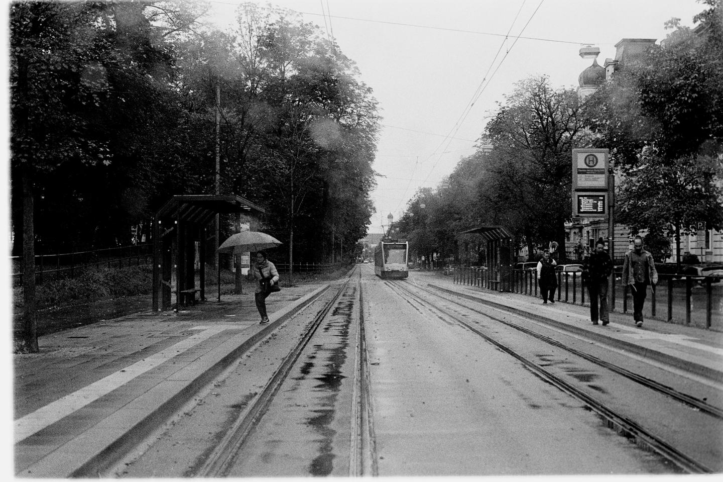 A woman with an umbrella stands at a tram stop while a tram approaches in the distance and people walk on the sidewalk.