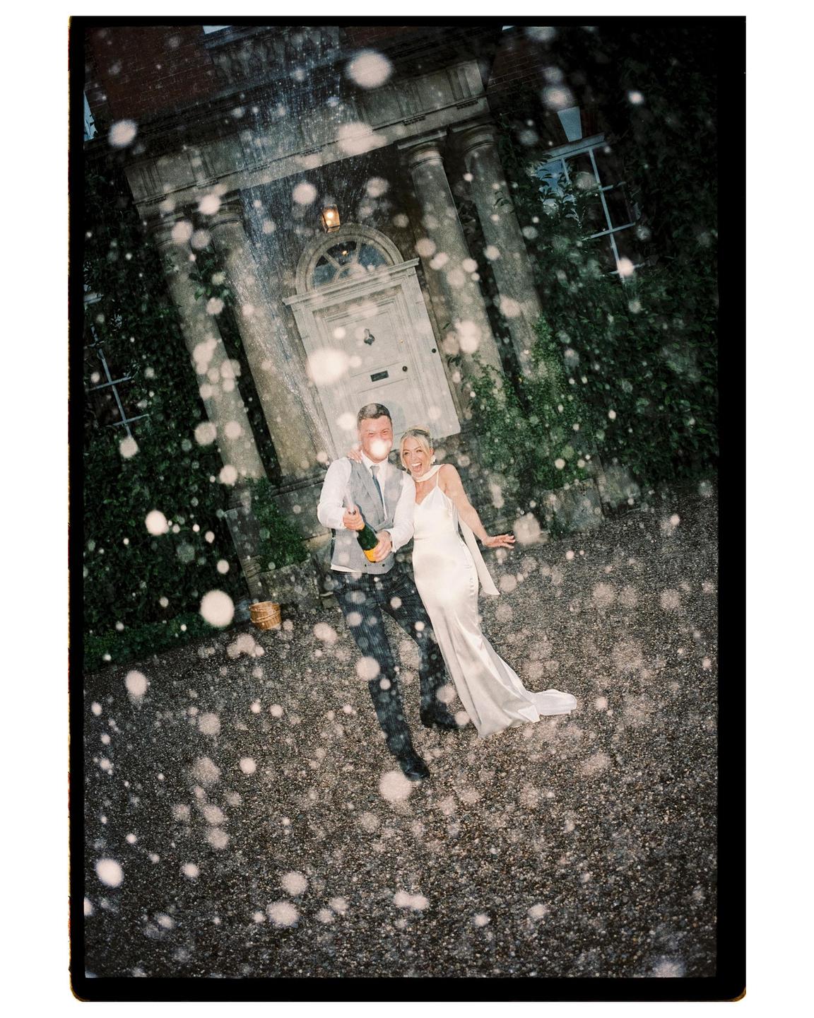 A couple stands in front of a large house with champagne spraying around them.