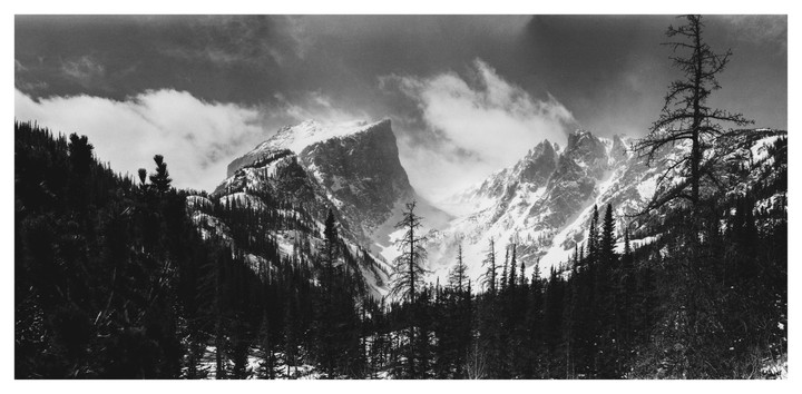A panoramic black and white landscape shows a snow-capped mountain peak surrounded by clouds, with a forest of pine trees in the foreground.