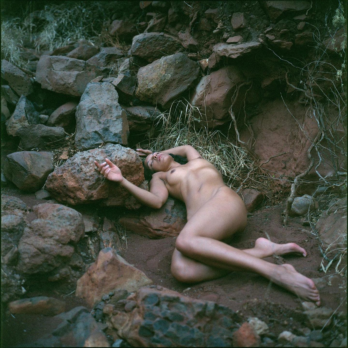 A nude woman lies on the ground among rocks and dry grass.