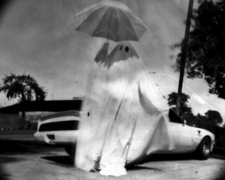 A person dressed as a ghost holds an umbrella while standing next to a car.