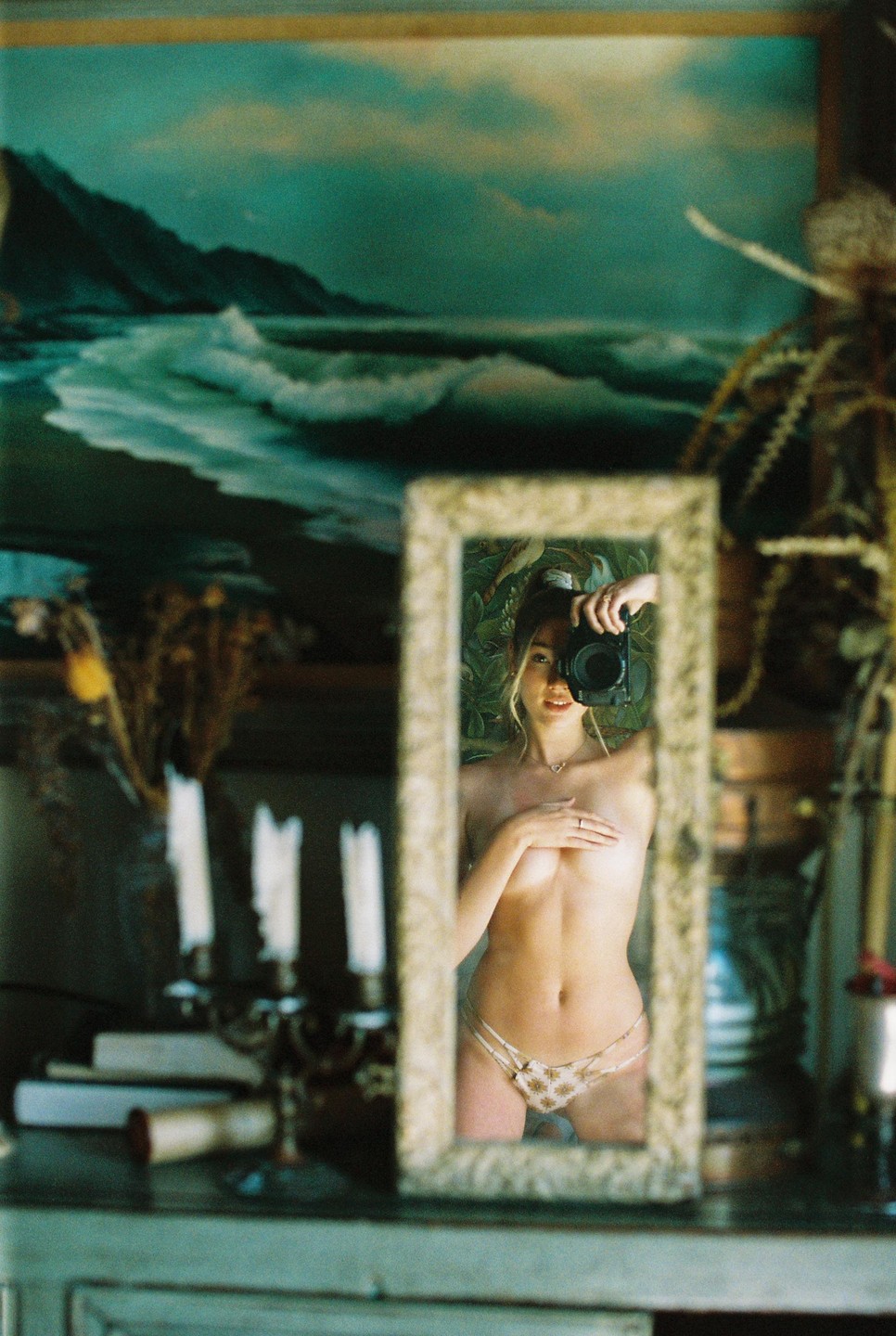 A woman takes a self-portrait in a mirror, with a painting of a seascape and a candelabra in the background.