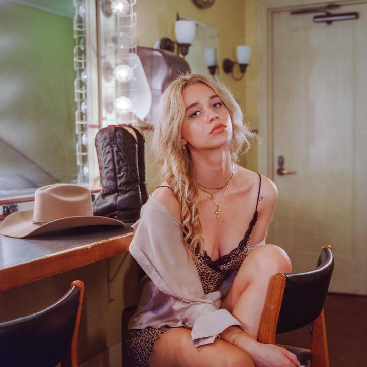 A woman with blonde hair sits on a chair next to a vanity with a cowboy hat and boots.