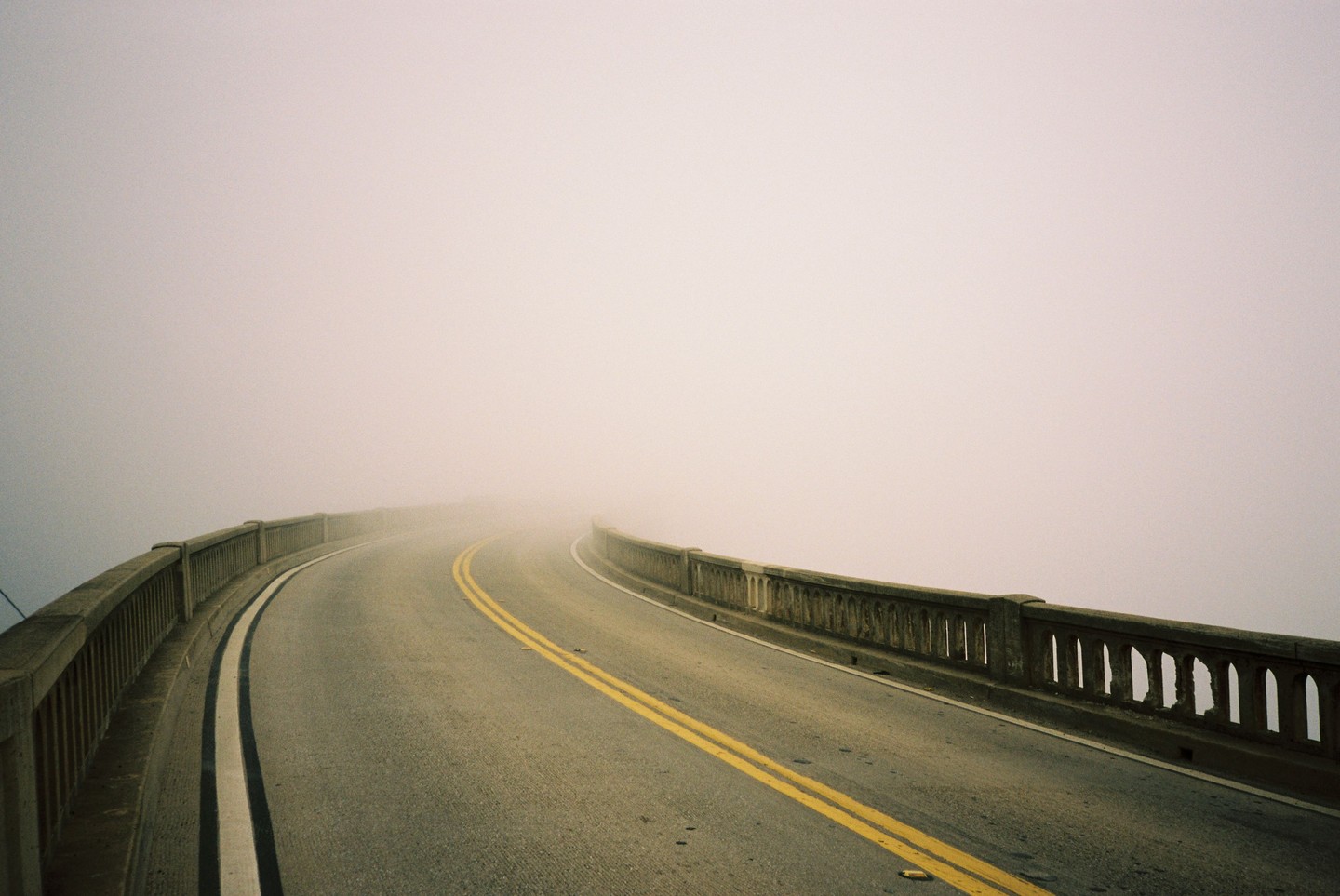 A winding road with a concrete barrier on both sides disappears into thick fog.