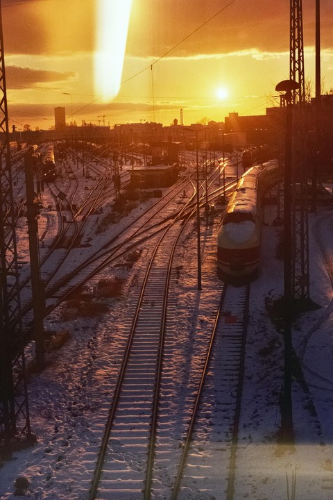 A train station with multiple tracks covered in snow is illuminated by the golden light of a sunset.