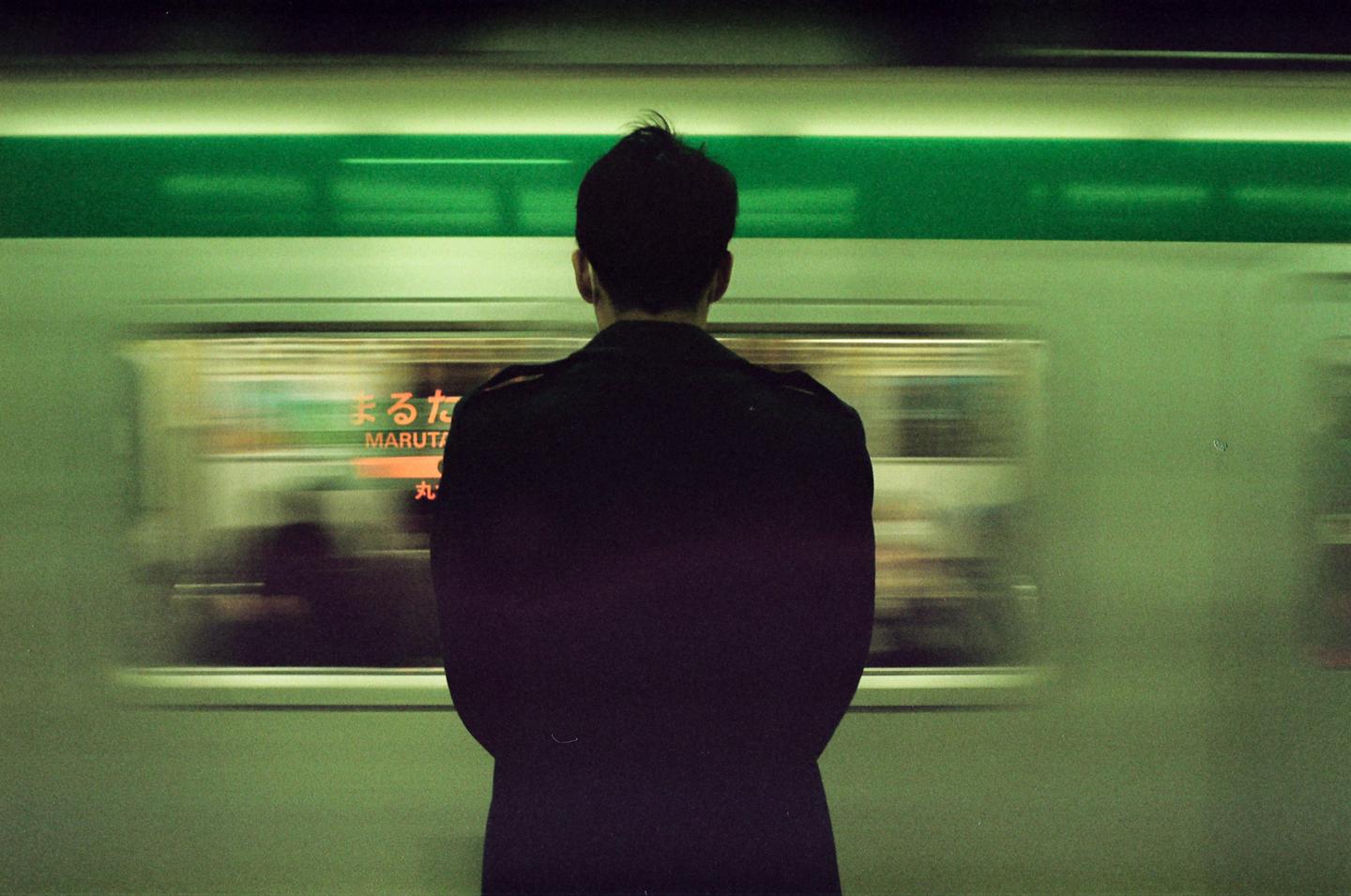 A man stands on a subway platform as a train passes by, creating a motion blur effect.
