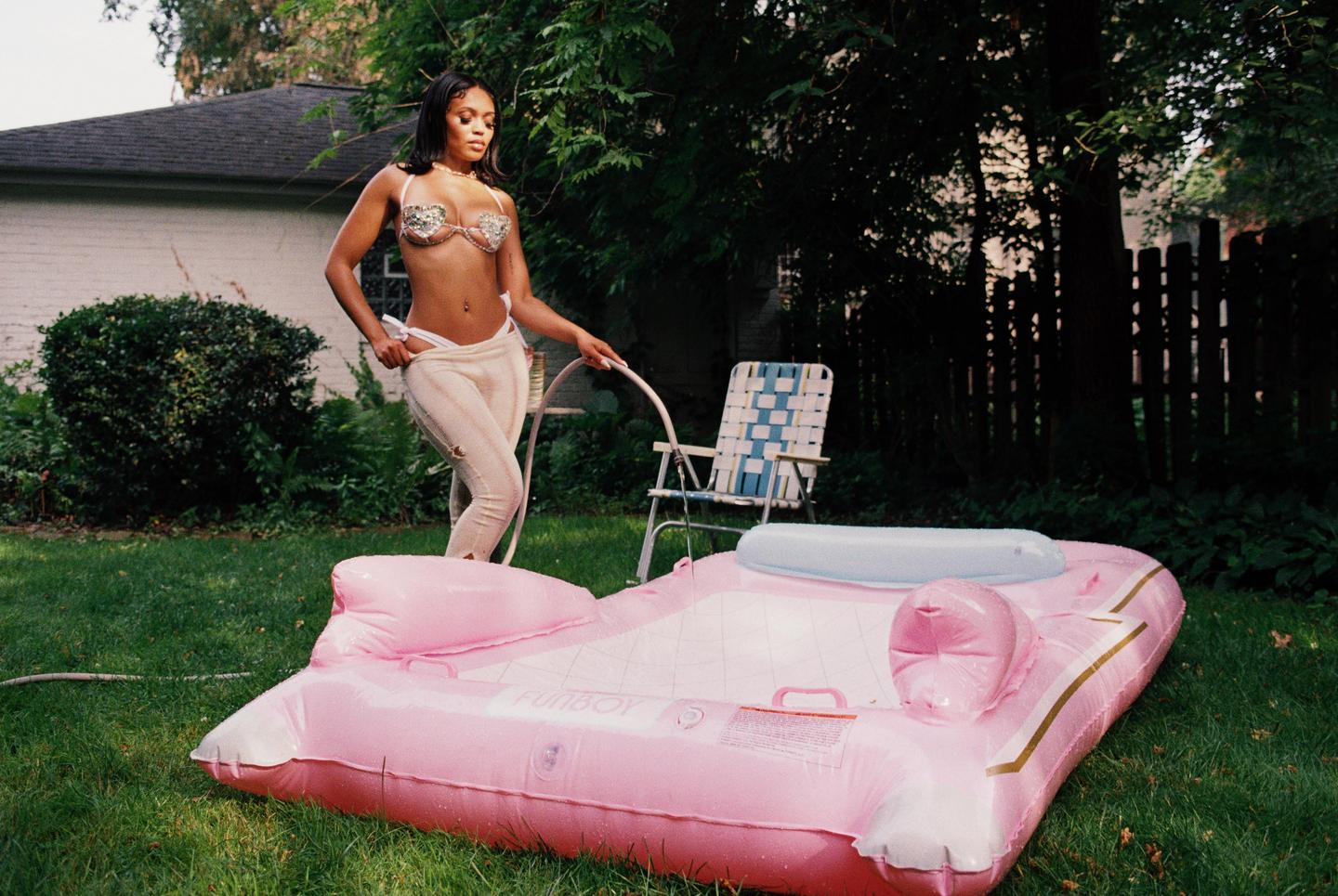 A woman in a sparkly bra and white pants fills a pink inflatable pool with a hose in a grassy backyard.