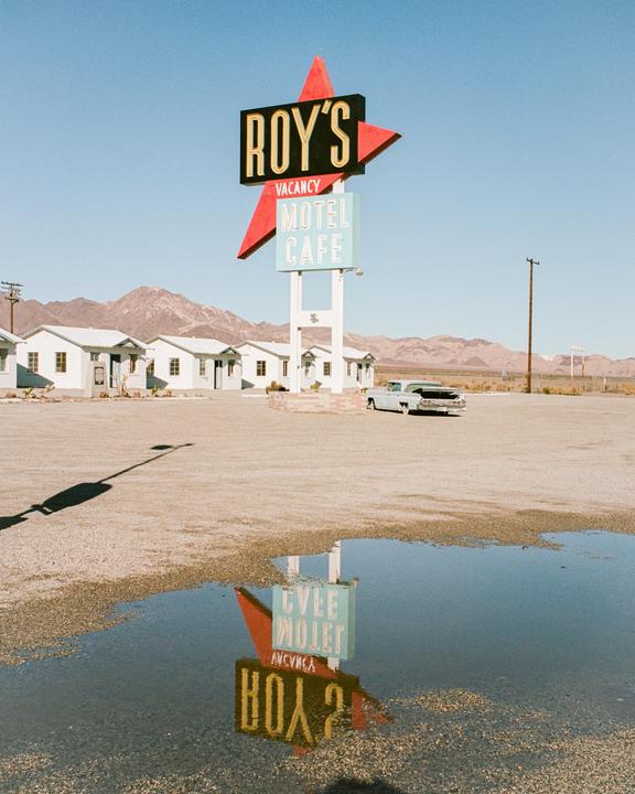 A motel sign is reflected in a puddle in front of a motel and mountains.