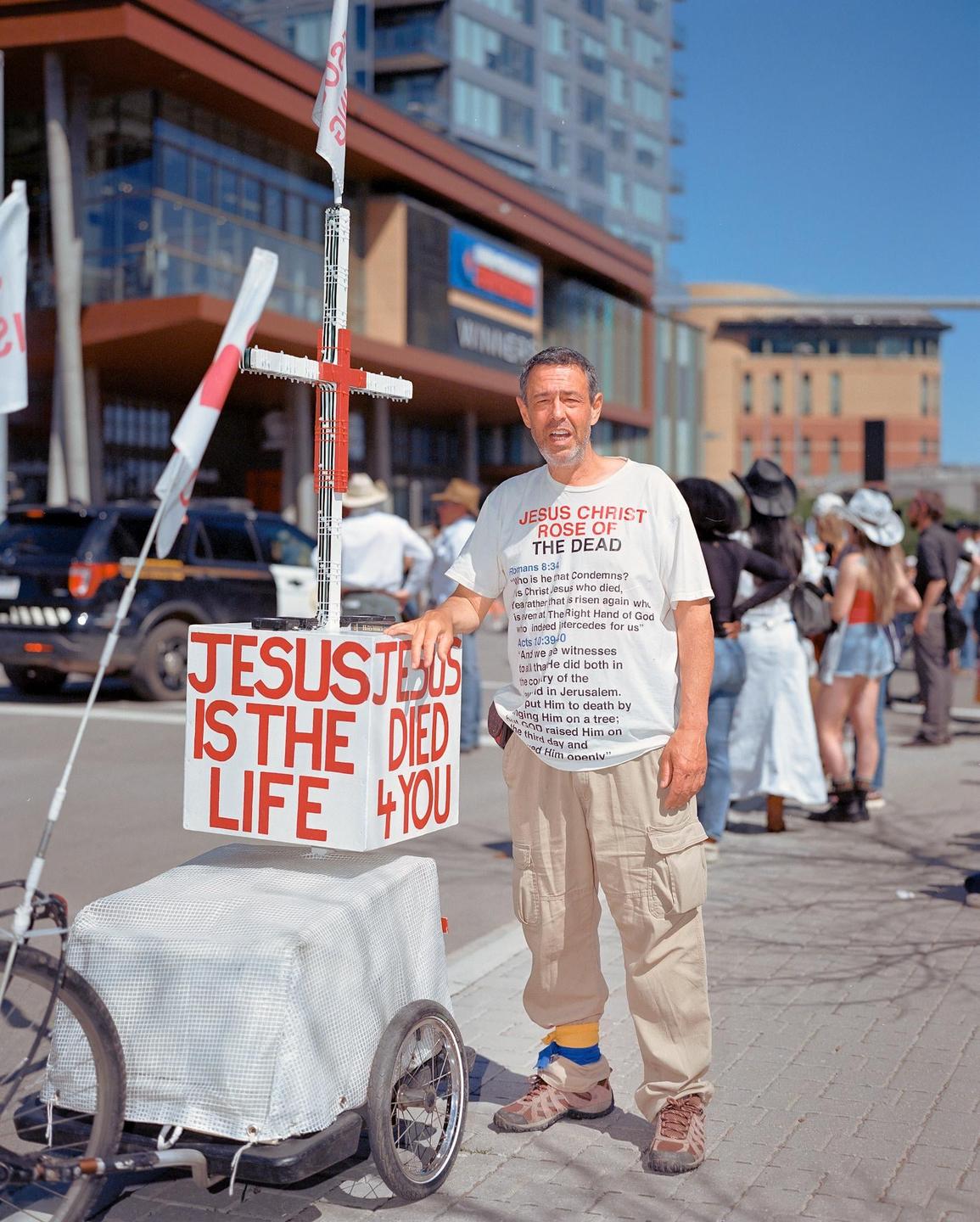 A man stands next to a cart with a cross and a sign that says "Jesus is the life 4 you" on a city street.