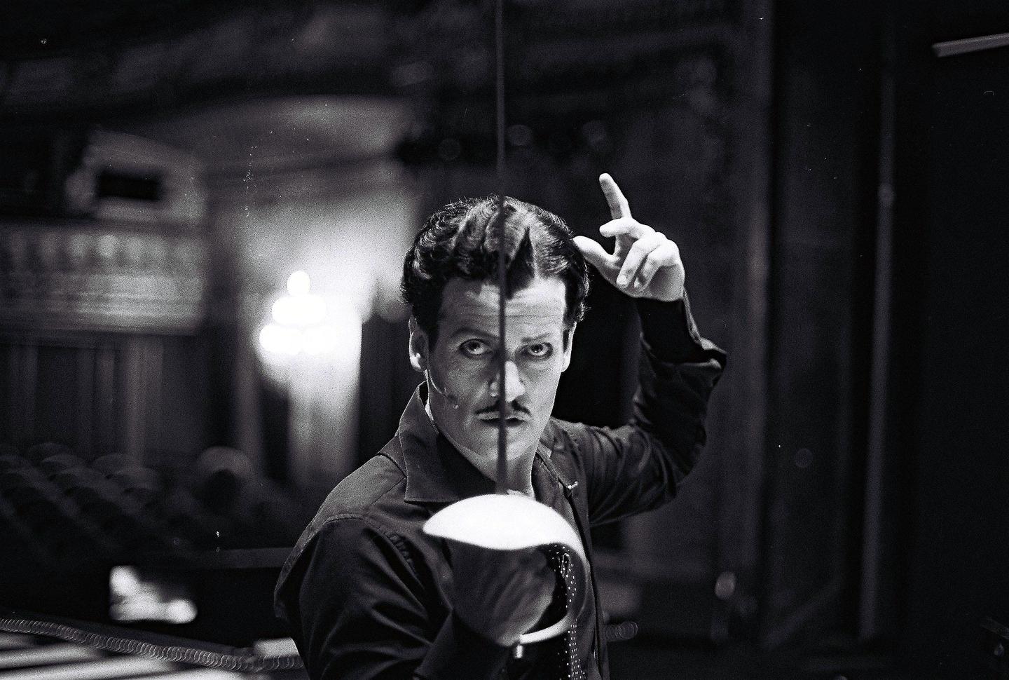 A man with a painted face and a mustache holds a fencing foil and points his finger up.