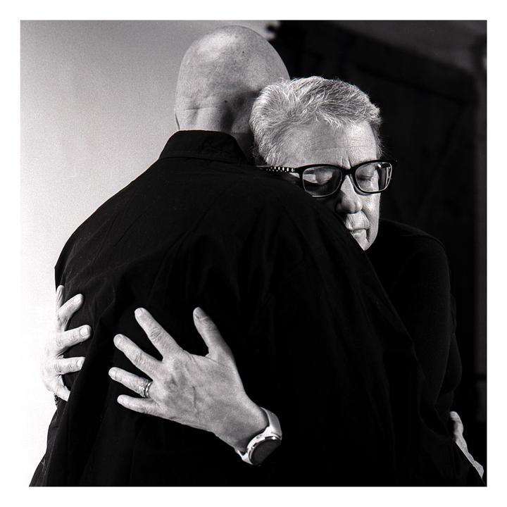 A man with glasses is embracing another man from behind.